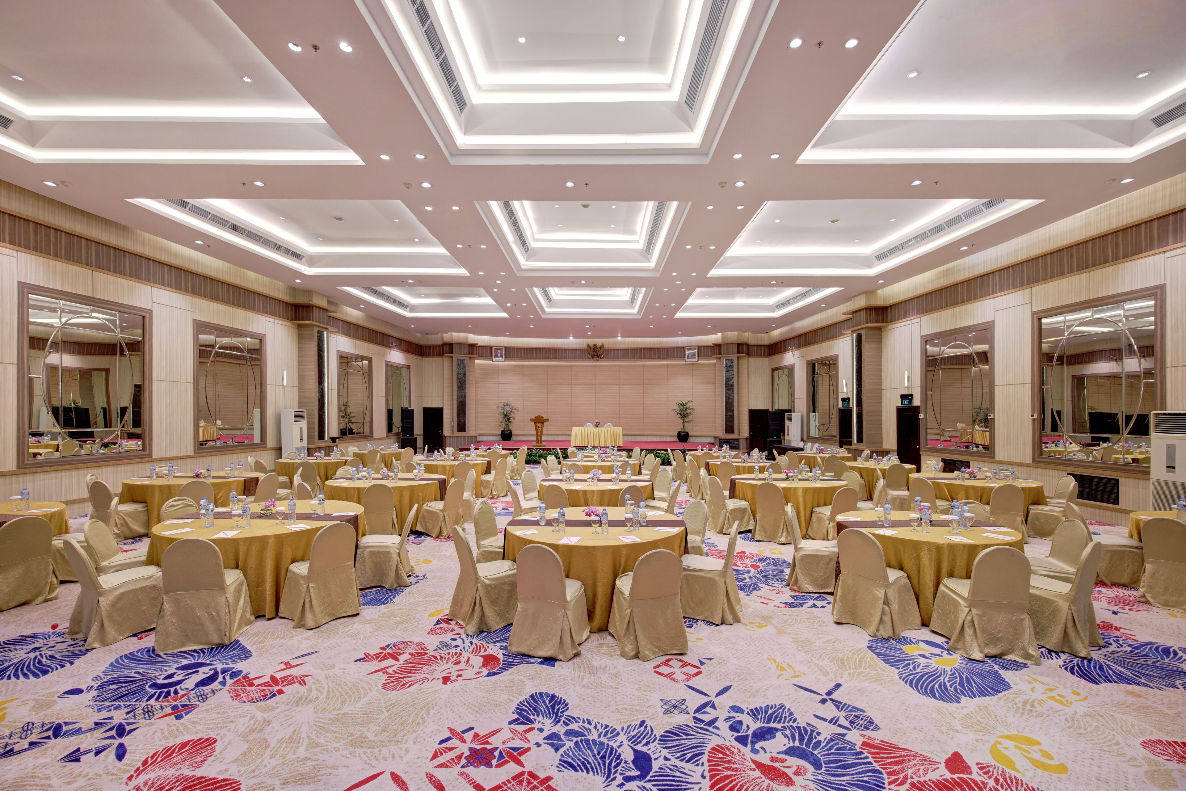 Un gran salón de banquetes con varias mesas redondas y sillas, una alfombra estampada y un techo alto con iluminación empotrada.