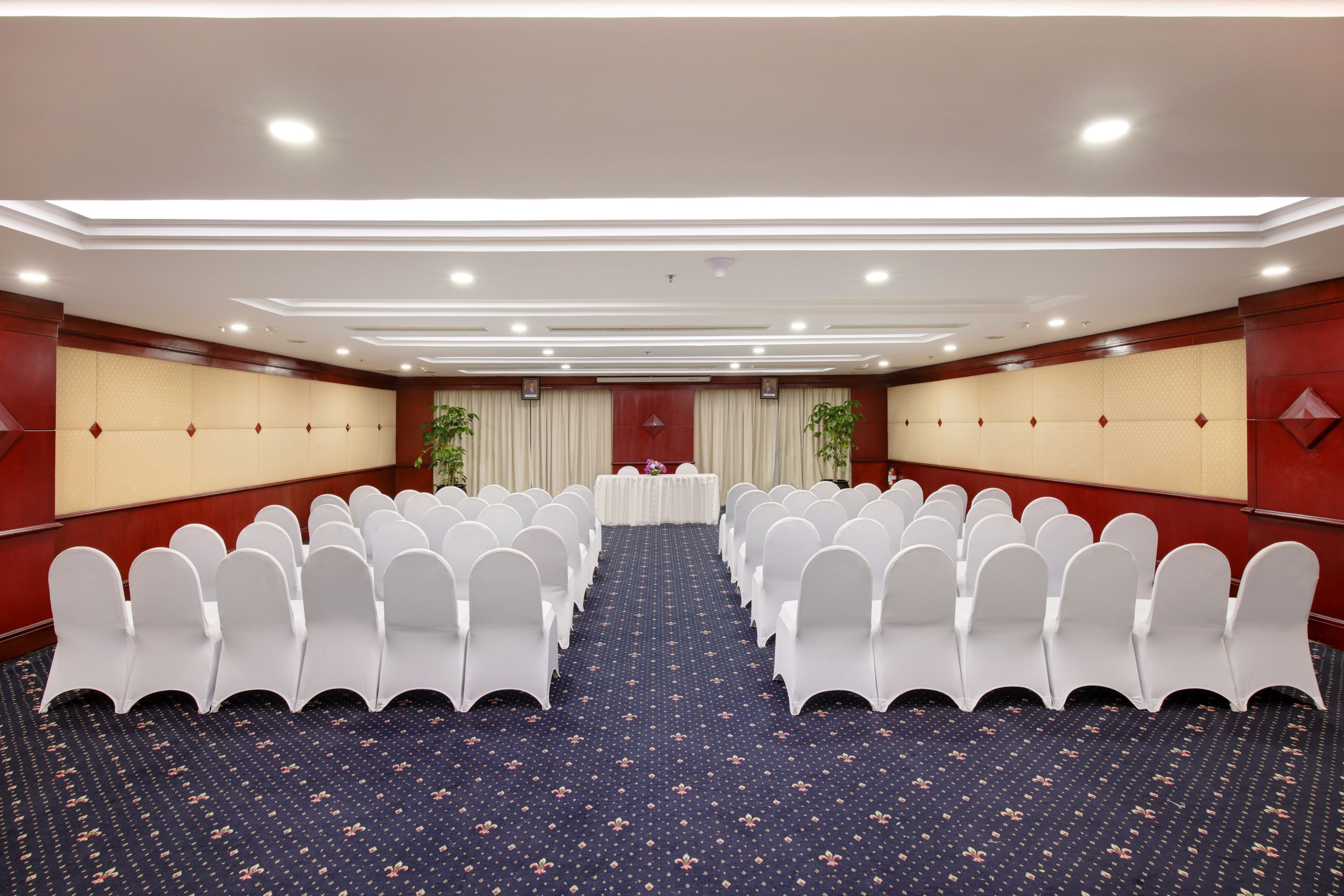Une salle de conférence avec des rangées de chaises blanches, une longue table et un tapis à motifs.