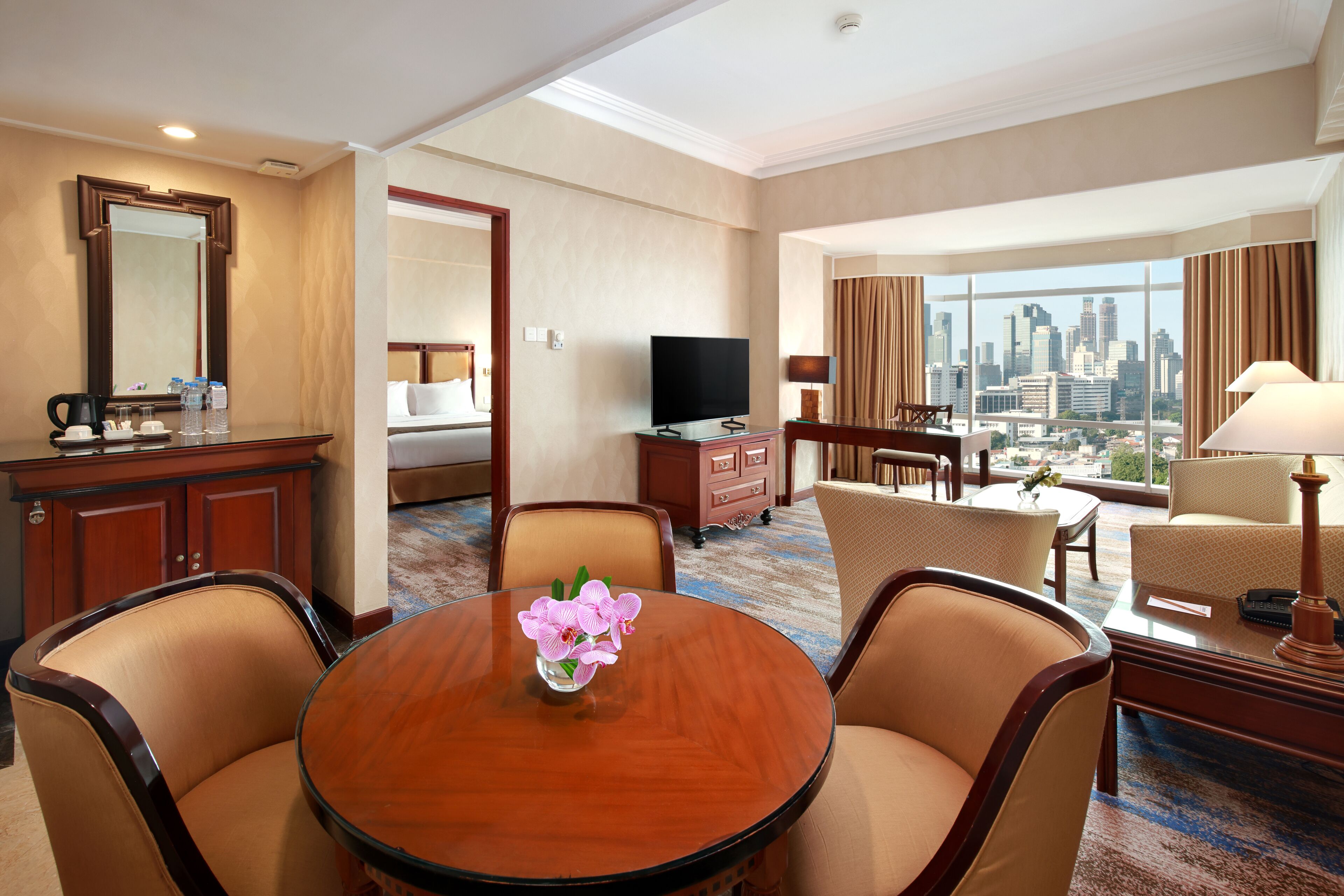 Habitación de hotel con vista a la ciudad, una mesa redonda de comedor con sillas, un televisor, un escritorio con lámpara y una cama con vista panorámica de la ciudad.
