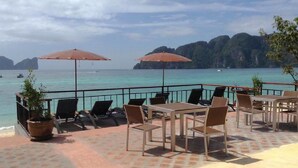 Una terraza frente al mar con mesas y sillas, dos sombrillas y vistas al océano y montañas lejanas.