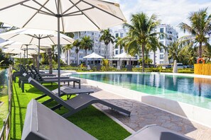 A poolside area with lounge chairs and umbrellas, a swimming pool, palm trees, and a modern building in the background. - Champa Island Nha Trang - Resort Hotel & Spa (Nha Trang)