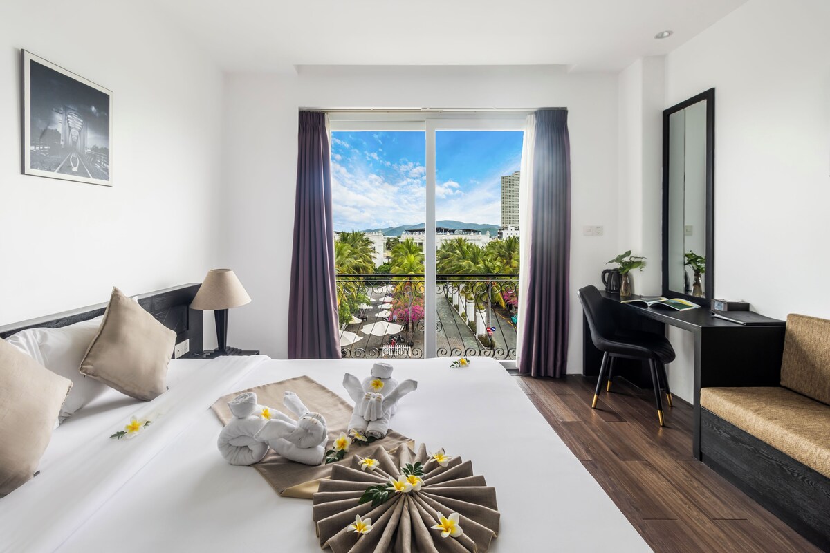 A hotel room with a bed, a desk, a chair, and a balcony with a view of palm trees and buildings.