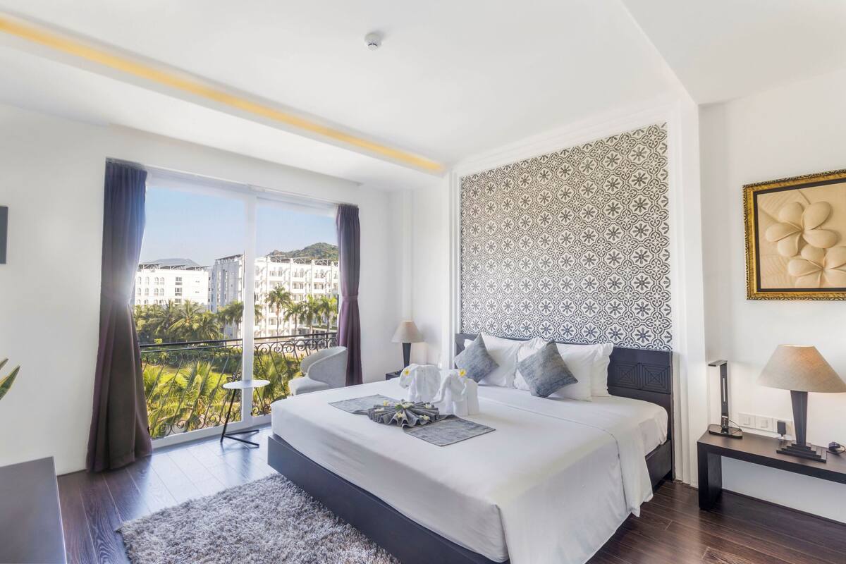 Une chambre d’hôtel moderne avec un grand lit, un balcon offrant une vue sur les palmiers et les immeubles, un mur décoré de motifs et une œuvre d’art encadrée.
