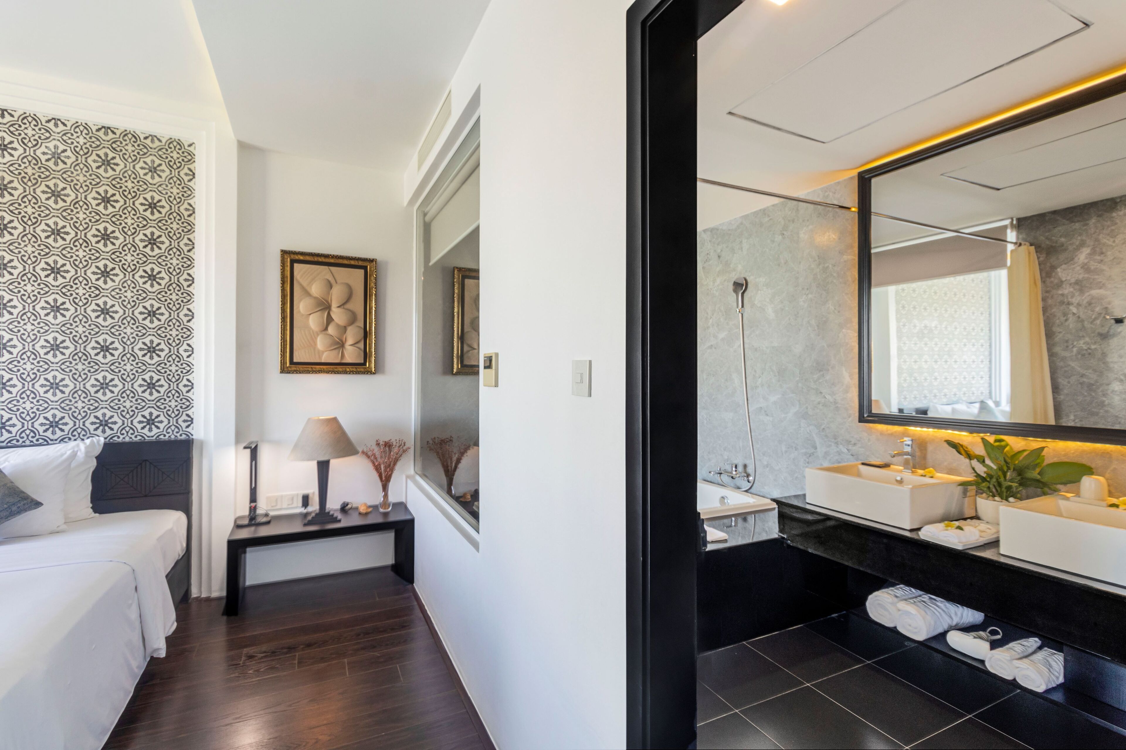 A modern hotel room with a bed, bedside table, lamp, framed artwork, and a bathroom with a large mirror, two sinks, and a shower.