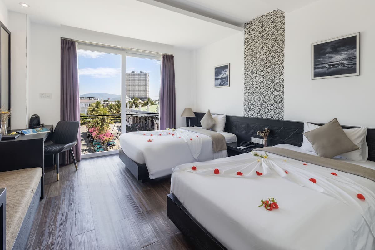 Une chambre d’hôtel avec deux lits, un bureau et un balcon offrant une vue sur les immeubles et les palmiers.