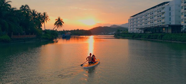 A river with a sunset, a boat with two people, and a building on the right. - Champa Island Nha Trang - Resort Hotel & Spa (Nha Trang)