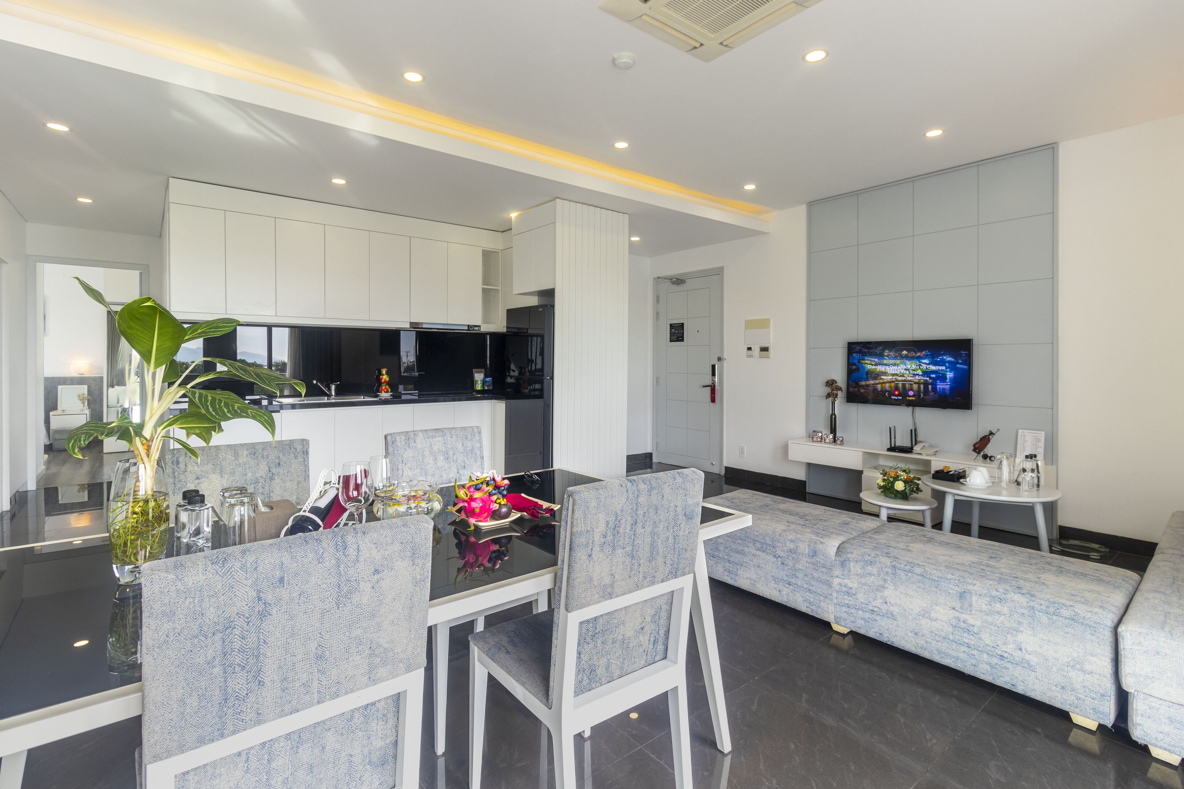 A modern dining area with a glass-top table, grey upholstered chairs, and a sofa. A flat-screen TV is mounted on the wall above the sofa.