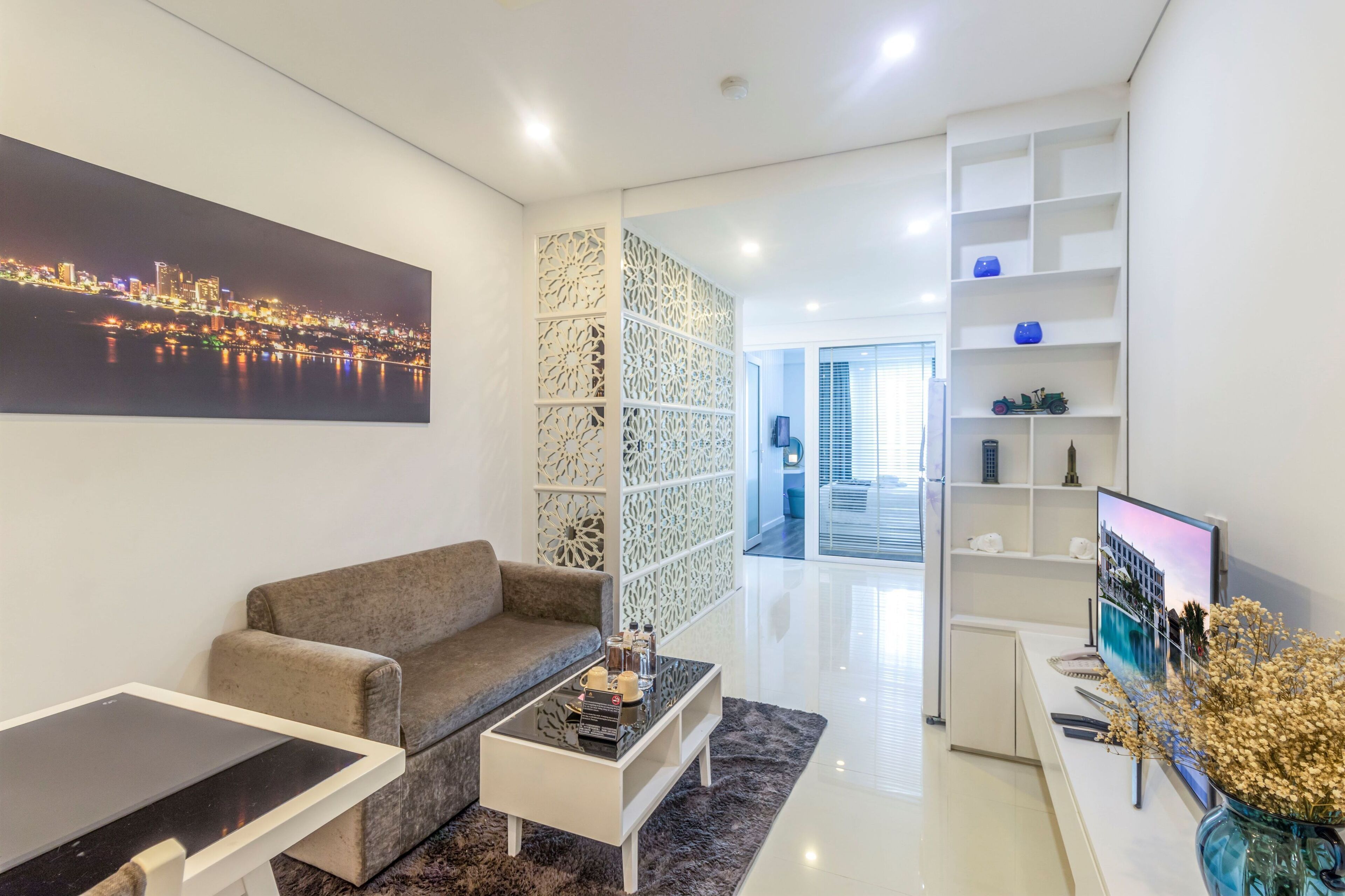 Een moderne woonkamer met een bank, salontafel en een televisie op een standaard. Er is een decoratief wandpaneel en een boekenkast met diverse spullen.