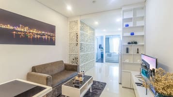 Een moderne woonkamer met een bank, salontafel en een televisie op een standaard. Er is een decoratief wandpaneel en een boekenkast met diverse spullen.