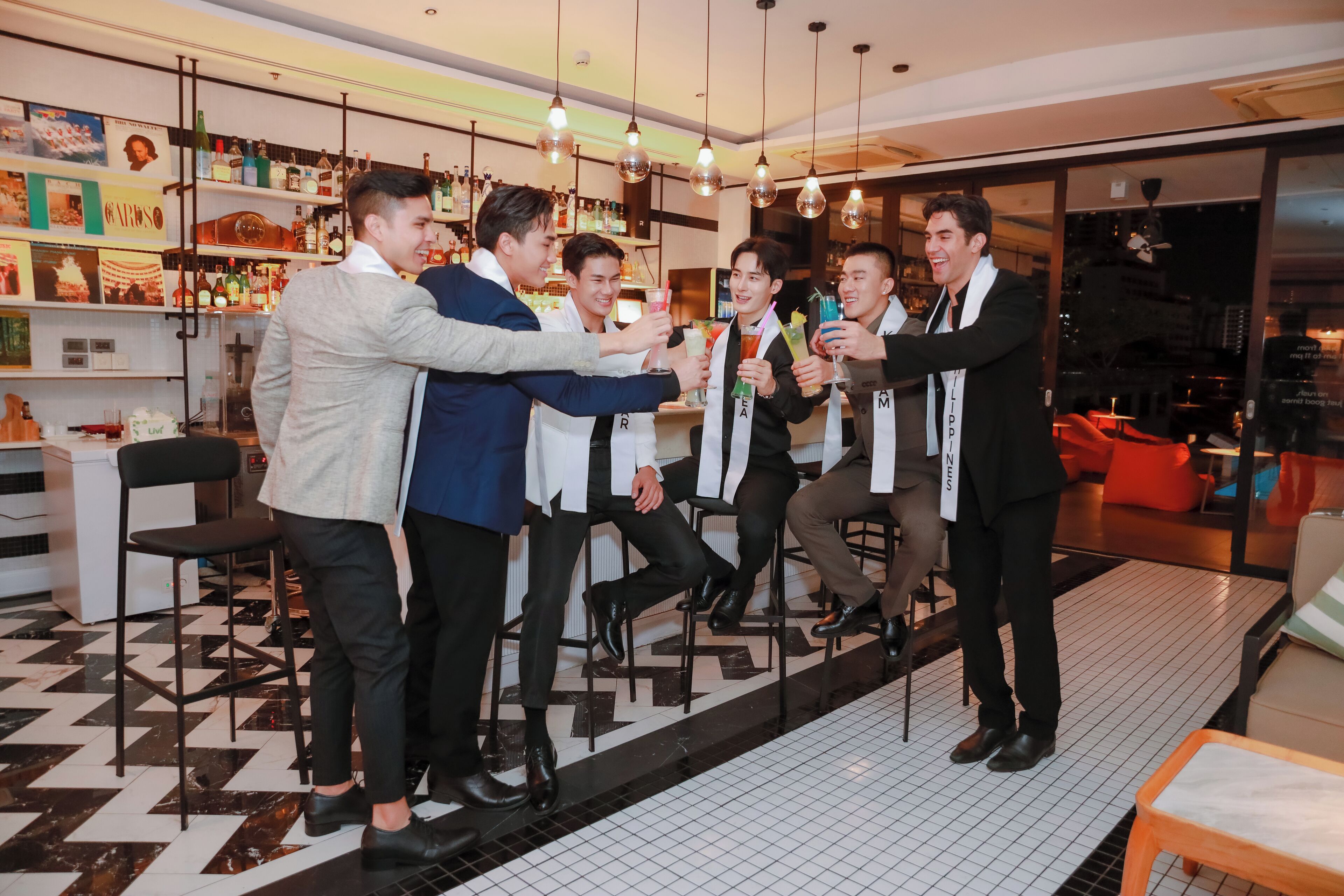 A group of men in suits toasting with drinks in a modern bar with tiled flooring and shelves of bottles.