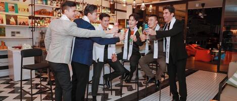 A group of men in suits toasting with drinks in a modern bar with tiled flooring and shelves of bottles.