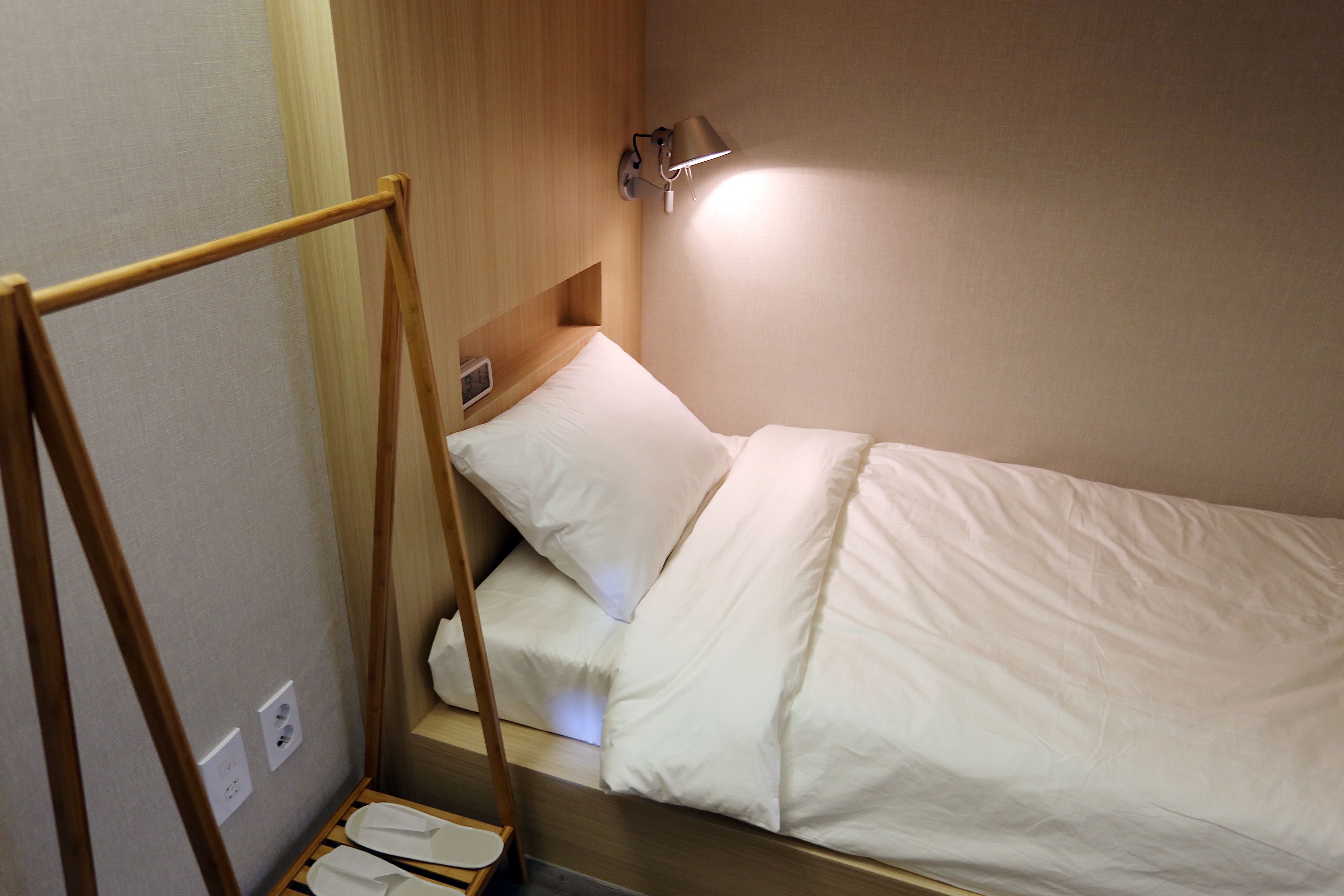 A small, neatly made bed with white bedding and a wooden headboard. There is a wall-mounted lamp providing light, and a wooden ladder attached to the headboard.