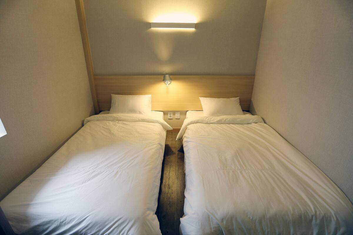 A small, narrow hotel room with two single beds, a wooden headboard, and a wall-mounted light fixture.