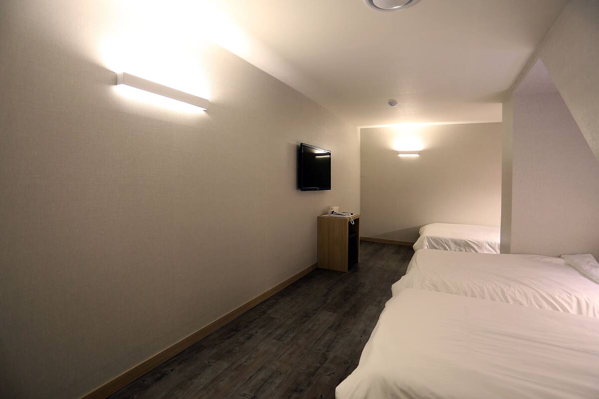 A hotel room with a wooden floor, a bed, a nightstand, a TV mounted on the wall, and a light fixture on the ceiling.