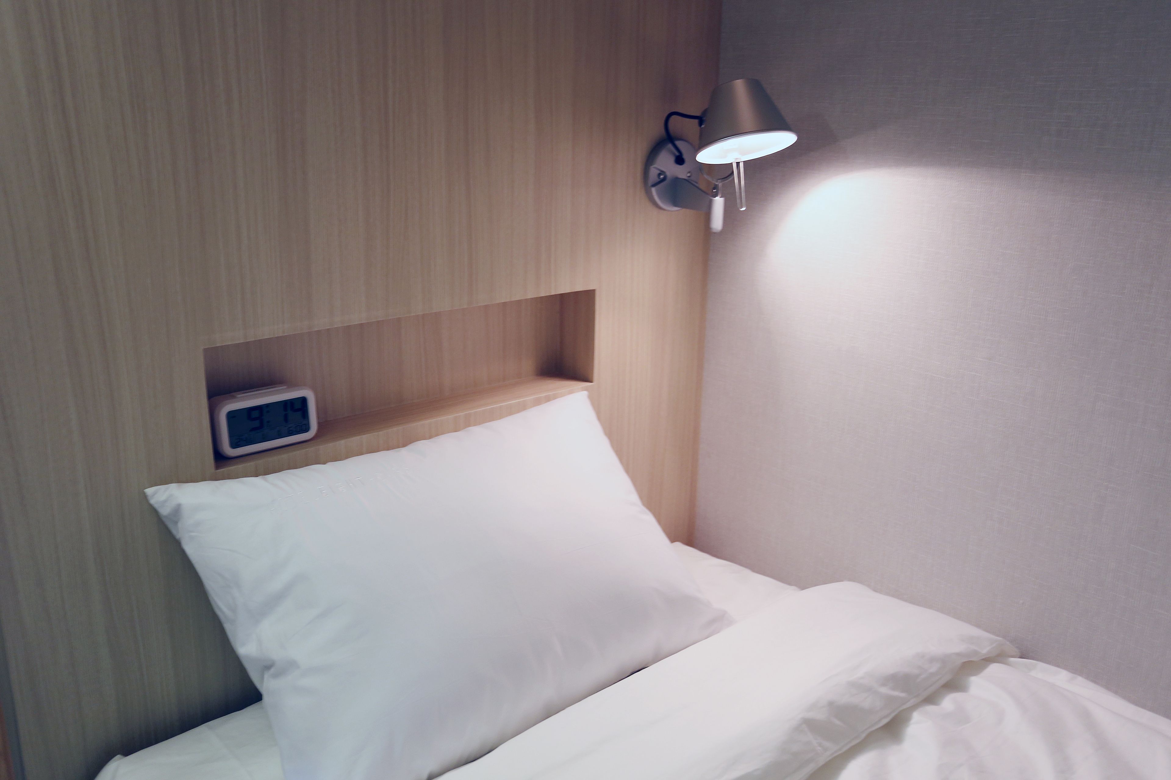 A neatly made bed with white bedding, a wall-mounted nightstand with a clock, and a wall-mounted lamp.