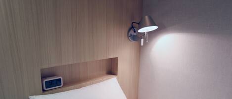 A neatly made bed with white bedding, a wall-mounted nightstand with a clock, and a wall-mounted lamp.