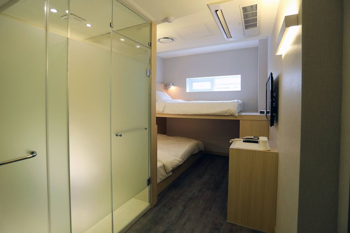 A compact room with a bunk bed, a small desk, a bedside table, and a window providing natural light.