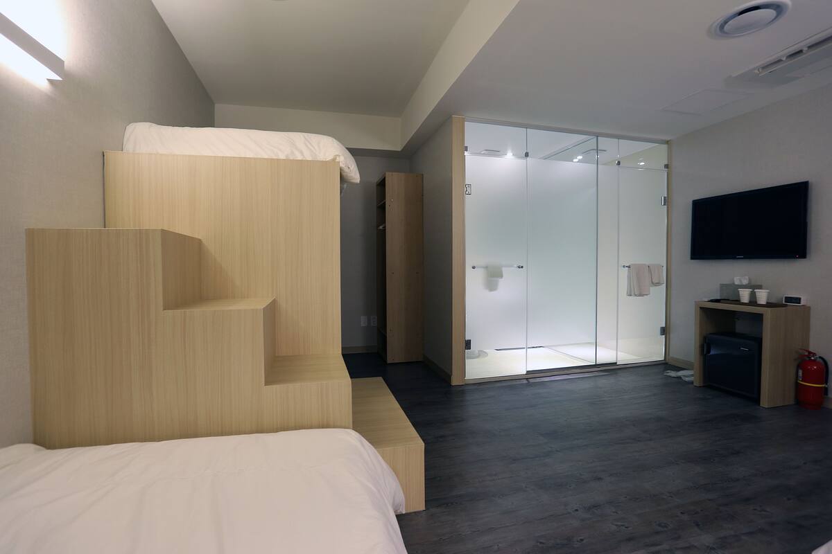 A modern hotel room with a bunk bed, a walk-in closet, a glass shower enclosure, a television, and a small desk with a fire extinguisher.