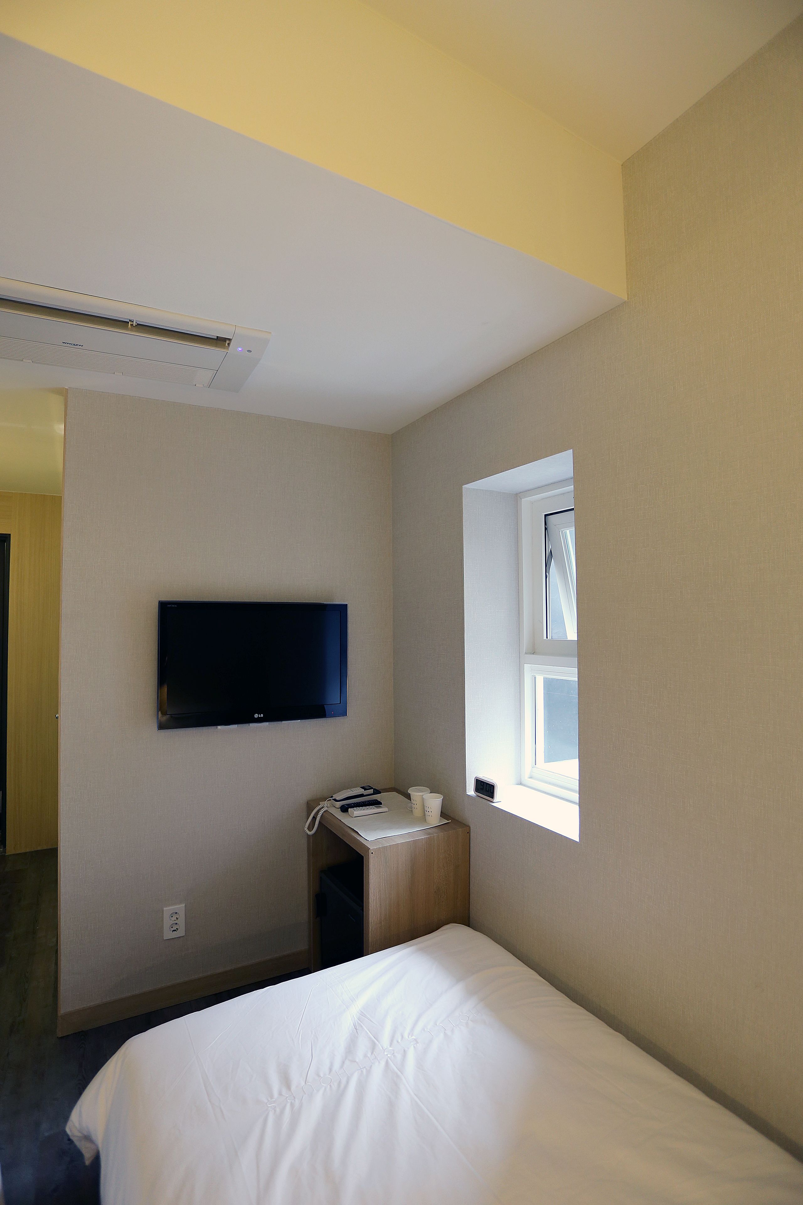 Ein Hotelzimmer mit einem Bett, einem an der Wand befestigten Fernseher, einem Fenster und einem kleinen Nachttisch mit einer Lampe.