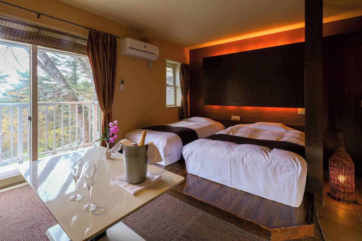 Habitación de hotel con dos camas, una mesa de comedor con dos copas de vino, un florero con flores y vista a árboles por una ventana.