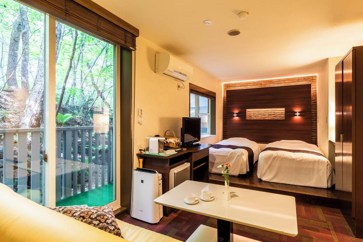 Habitación de hotel con balcón, escritorio con televisor, dos camas y una pequeña mesa con tazas y un florero.