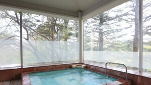 Un bain à remous rectangulaire avec un escalier en métal, entouré d’un sol carrelé et protégé par une paroi en verre offrant une vue sur les arbres.
