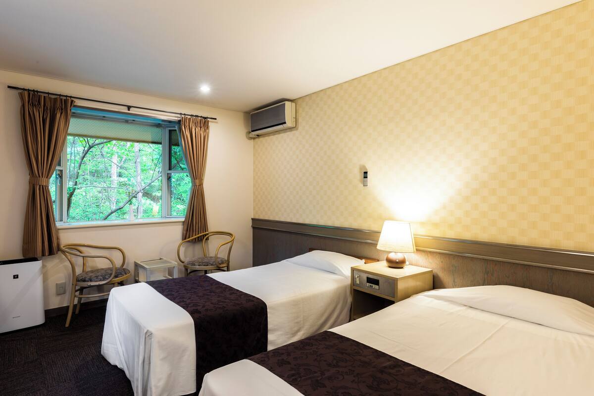Habitación de hotel con dos camas, una ventana con cortinas, un escritorio con lámpara y una silla.