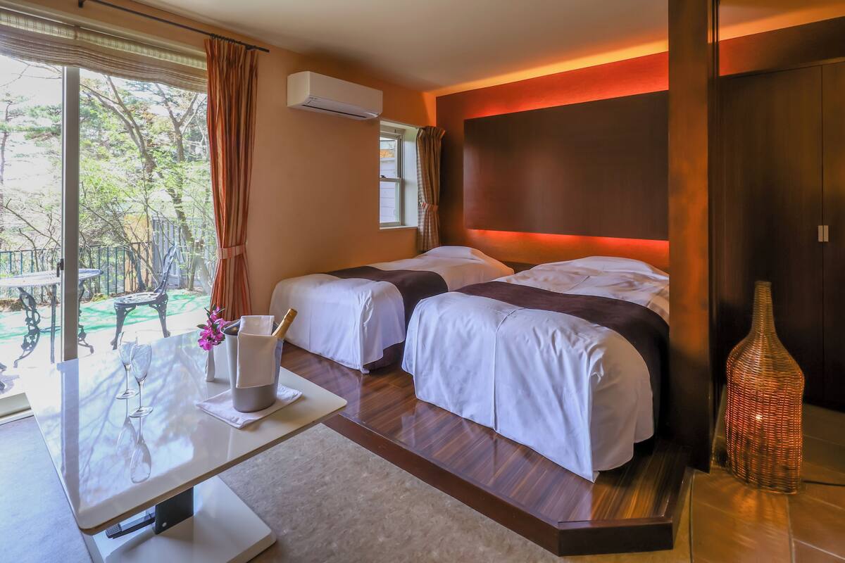 Una habitación de hotel con dos camas, una mesa con un florero y vasos, y vistas al exterior.