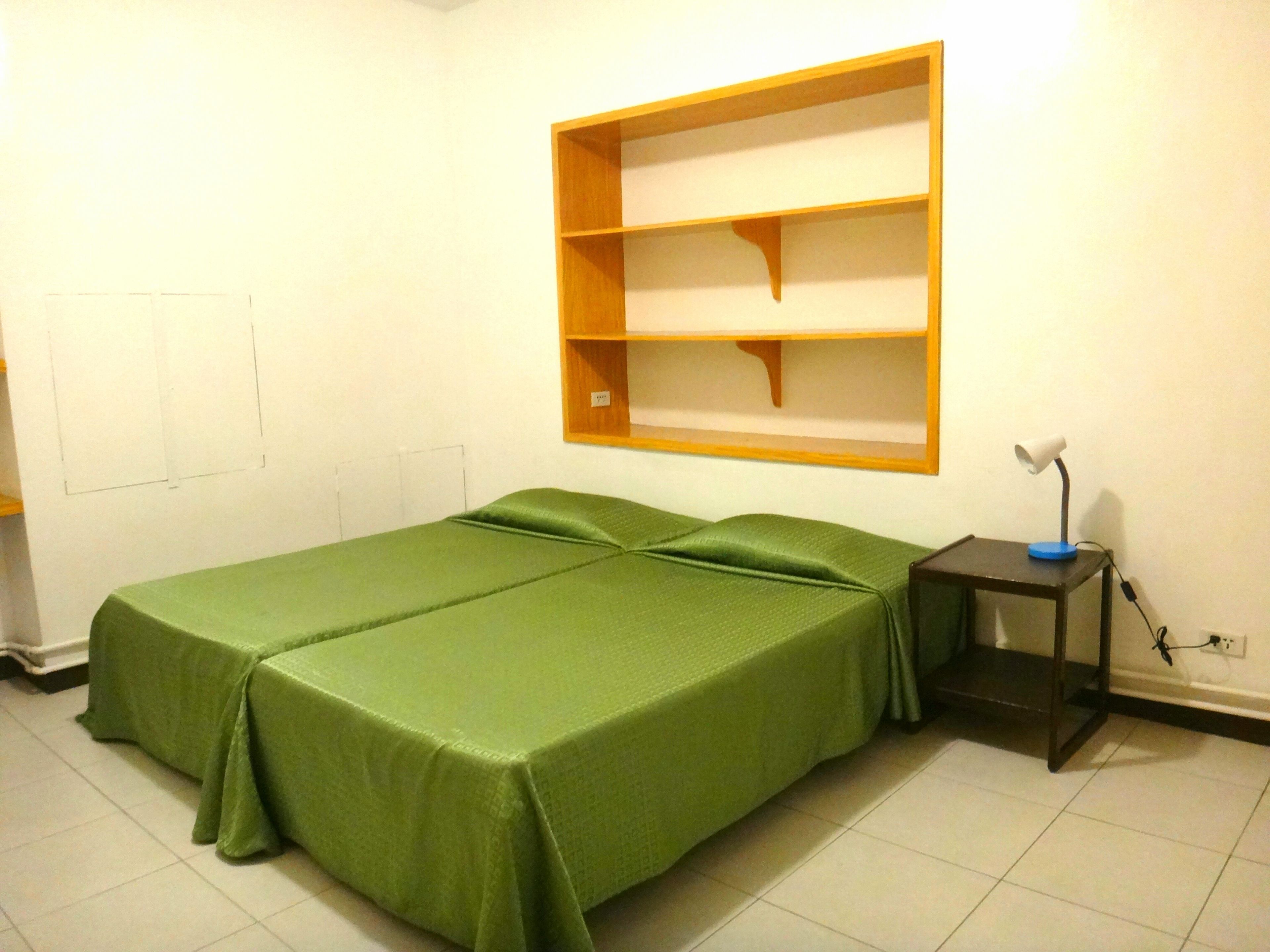 Une pièce avec un lit vert, une étagère en bois, un bureau avec une lampe et une prise de courant.