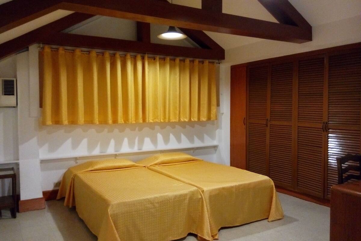 A room with a bed covered in yellow bedding, wooden beams on the ceiling, and a window with yellow curtains.
