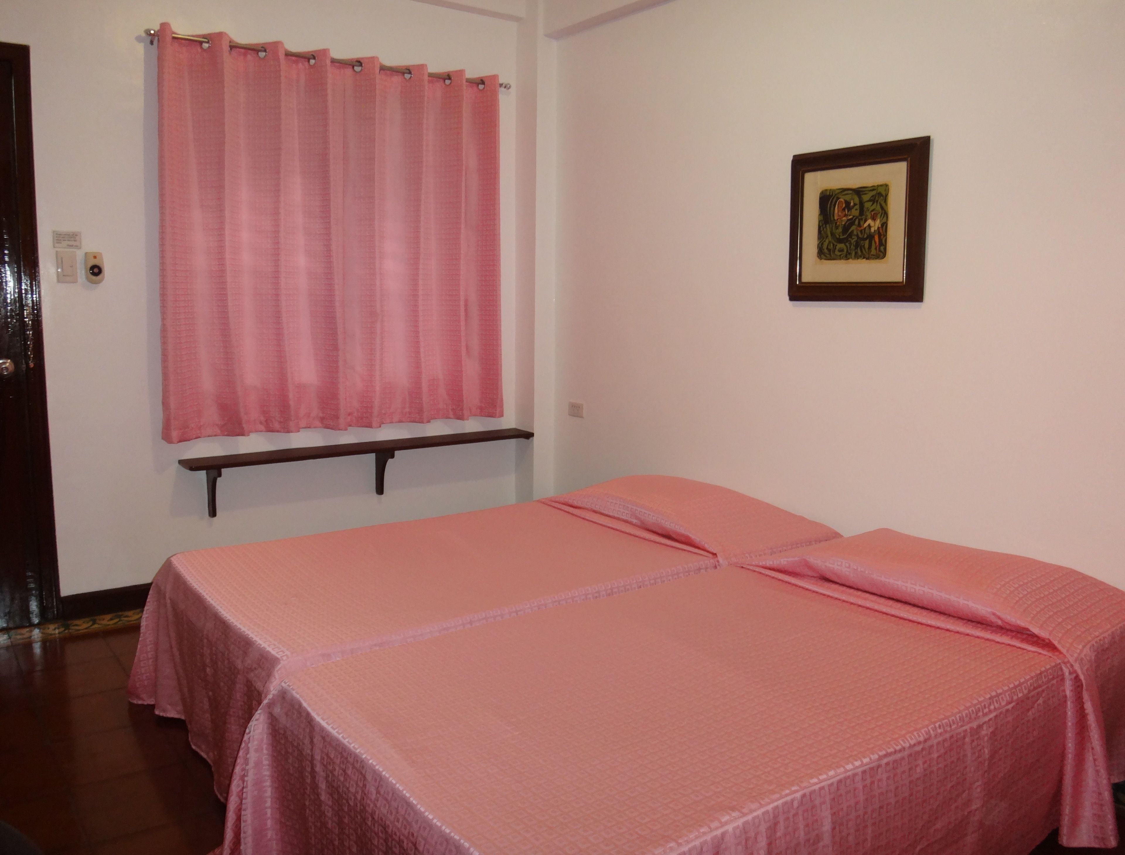 A room with two beds covered in pink bedding, a wooden bench, a pink curtain, and a framed picture on the wall.