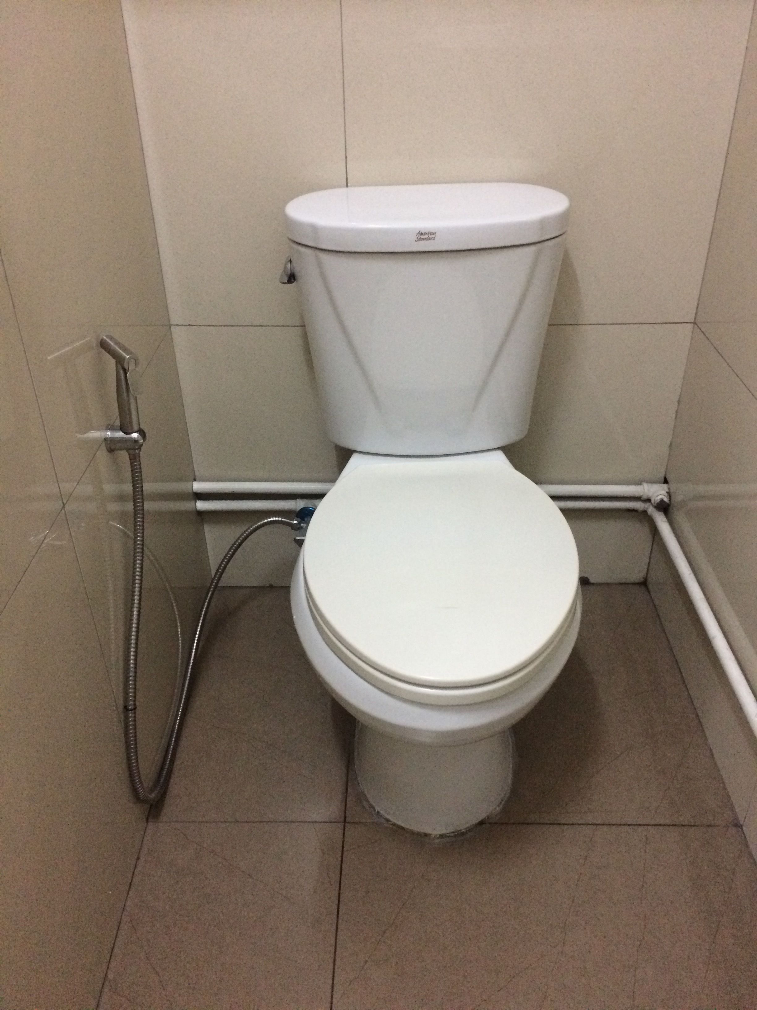 Inodoro blanco estándar con tanque y asiento, ubicado en un rincón de un baño con paredes y piso de baldosas color beige.