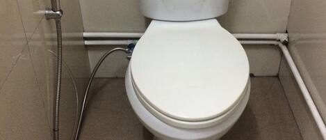 Inodoro blanco estándar con tanque y asiento, ubicado en un rincón de un baño con paredes y piso de baldosas color beige.