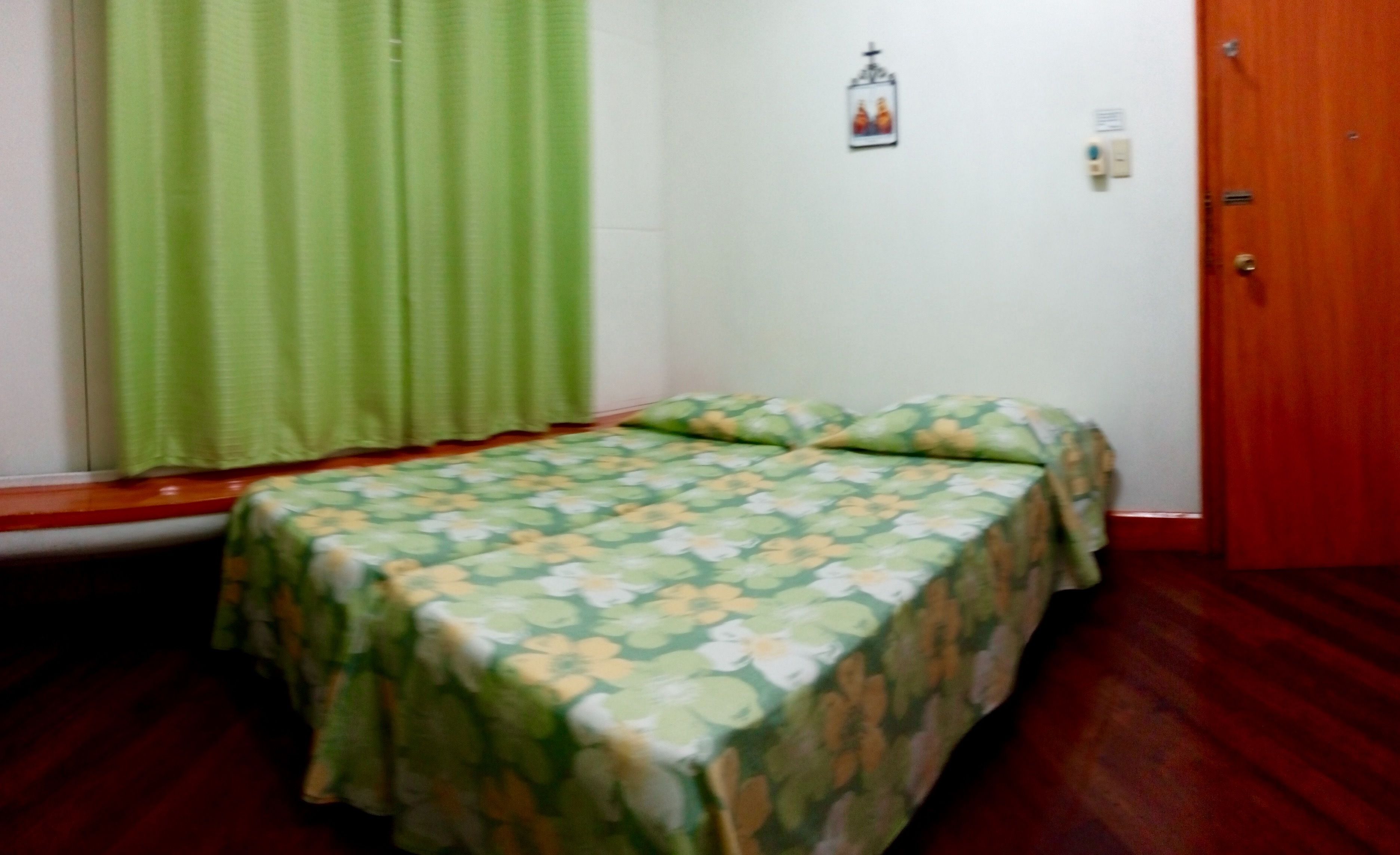 A room with a bed, green curtains, a wooden door, and a small wall-mounted light fixture.