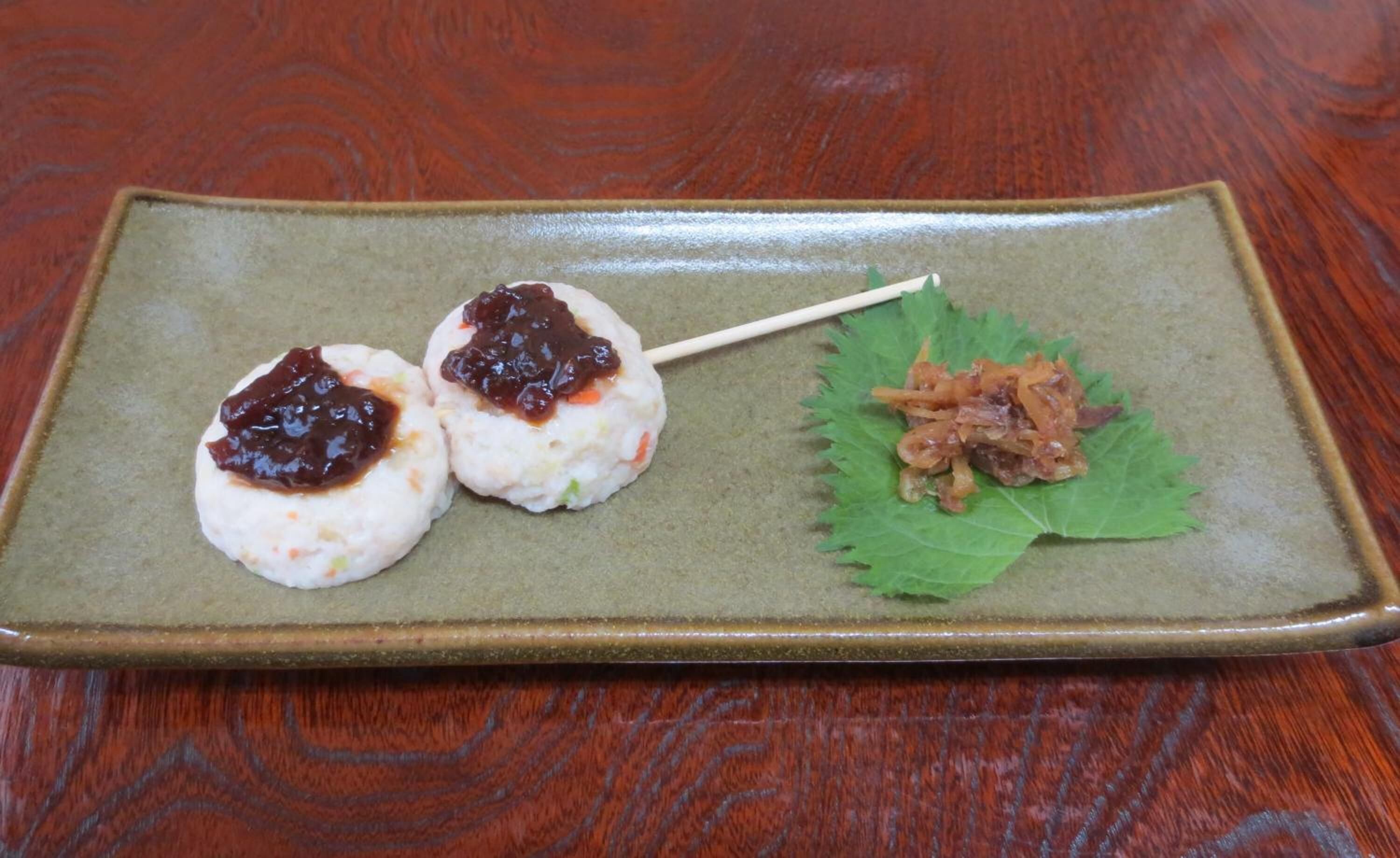 A rectangular ceramic plate with two dumplings topped with a dark sauce, and a side of pickled vegetables on a green leaf.