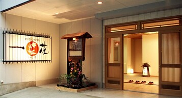 Una entrada tradicional japonesa con un portón de madera, un letrero con caracteres japoneses y un pequeño jardín con flores.