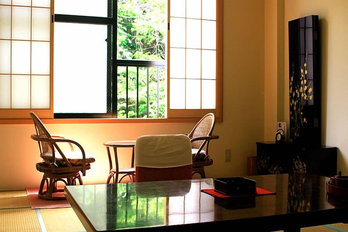 A traditional Japanese room with tatami flooring, a low table, two chairs, and a large window with a view of greenery.