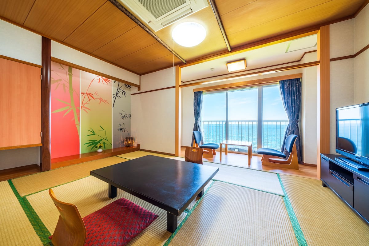 A room with tatami flooring, a low table, a sofa, a TV, and a view of the sea.