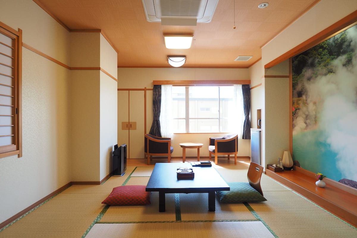 A traditional Japanese room with tatami flooring, a low table, two chairs, and a large window with curtains.