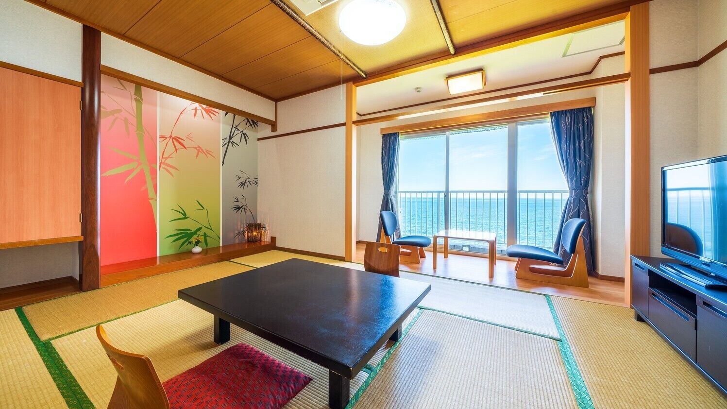 A room with tatami flooring, a low table, chairs, a television, and a large window with a view of the sea.