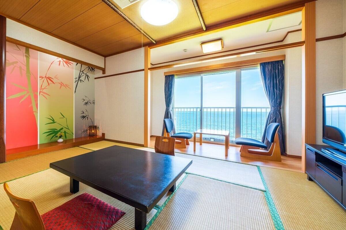 A room with tatami flooring, a low table, chairs, a television, and a large window with a view of the sea.