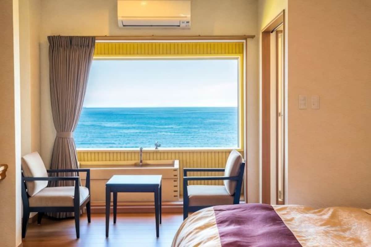 Ein Hotelzimmer mit Bett, zwei Stühlen, einem kleinen Tisch, einem Fenster mit Meerblick und einer Klimaanlage.