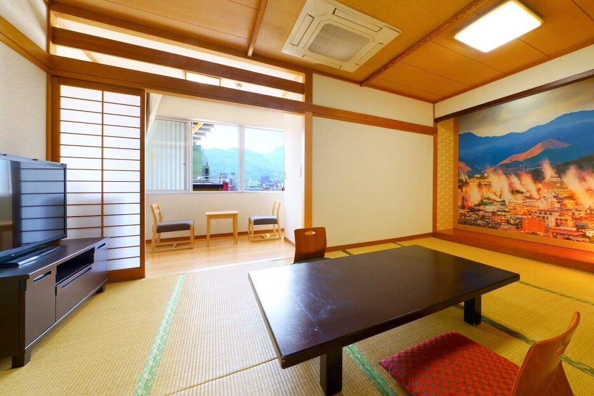 A traditional Japanese room with tatami flooring, a low table, chairs, a television, and a painting of a cityscape.
