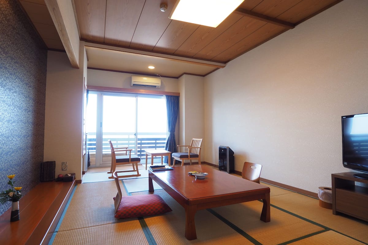A traditional Japanese room with tatami flooring, a wooden table, chairs, a television, and a sliding door leading to a balcony.