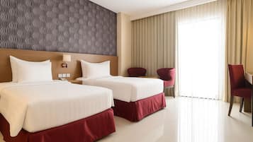 A hotel room with two beds, a headboard with a wavy pattern, a desk with a chair, and a large window with curtains.