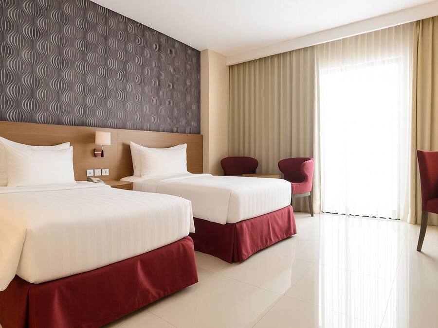 A hotel room with two beds, a headboard with a wavy pattern, a desk with a chair, and a large window with curtains.