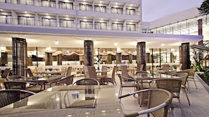 Un espace extérieur aménagé pour les repas, avec des meubles en osier et des tables en verre, entouré d’un bâtiment doté de nombreux balcons et d’une galerie couverte.
