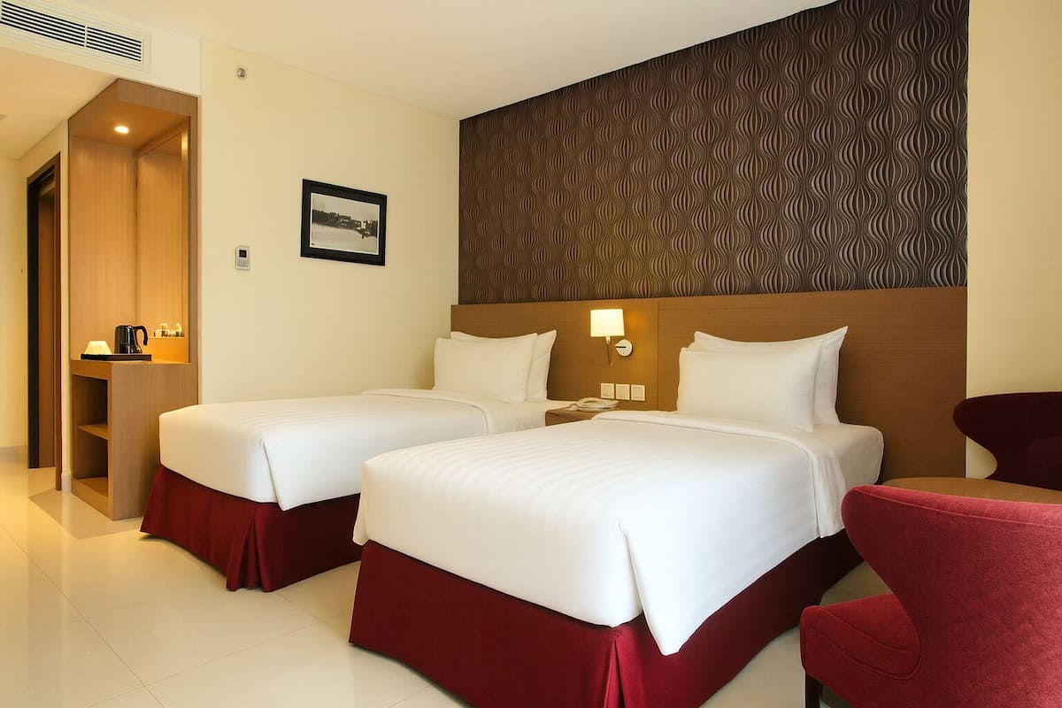 A hotel room with two beds, a wooden wardrobe, a small desk with a coffee maker, and a red armchair.