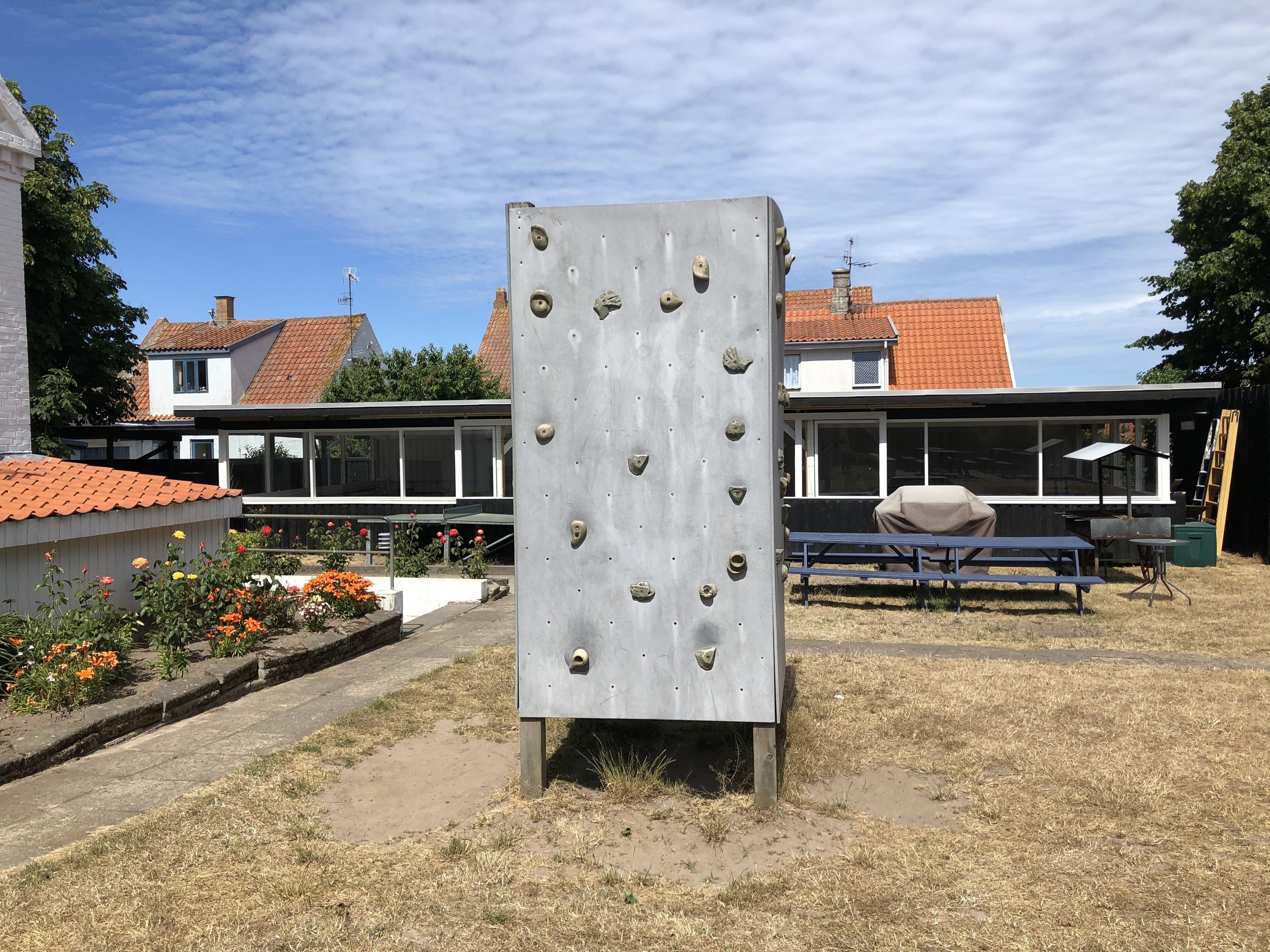 A climbing wall with a metal structure, a covered picnic table, and a bench in a garden area with houses in the background.