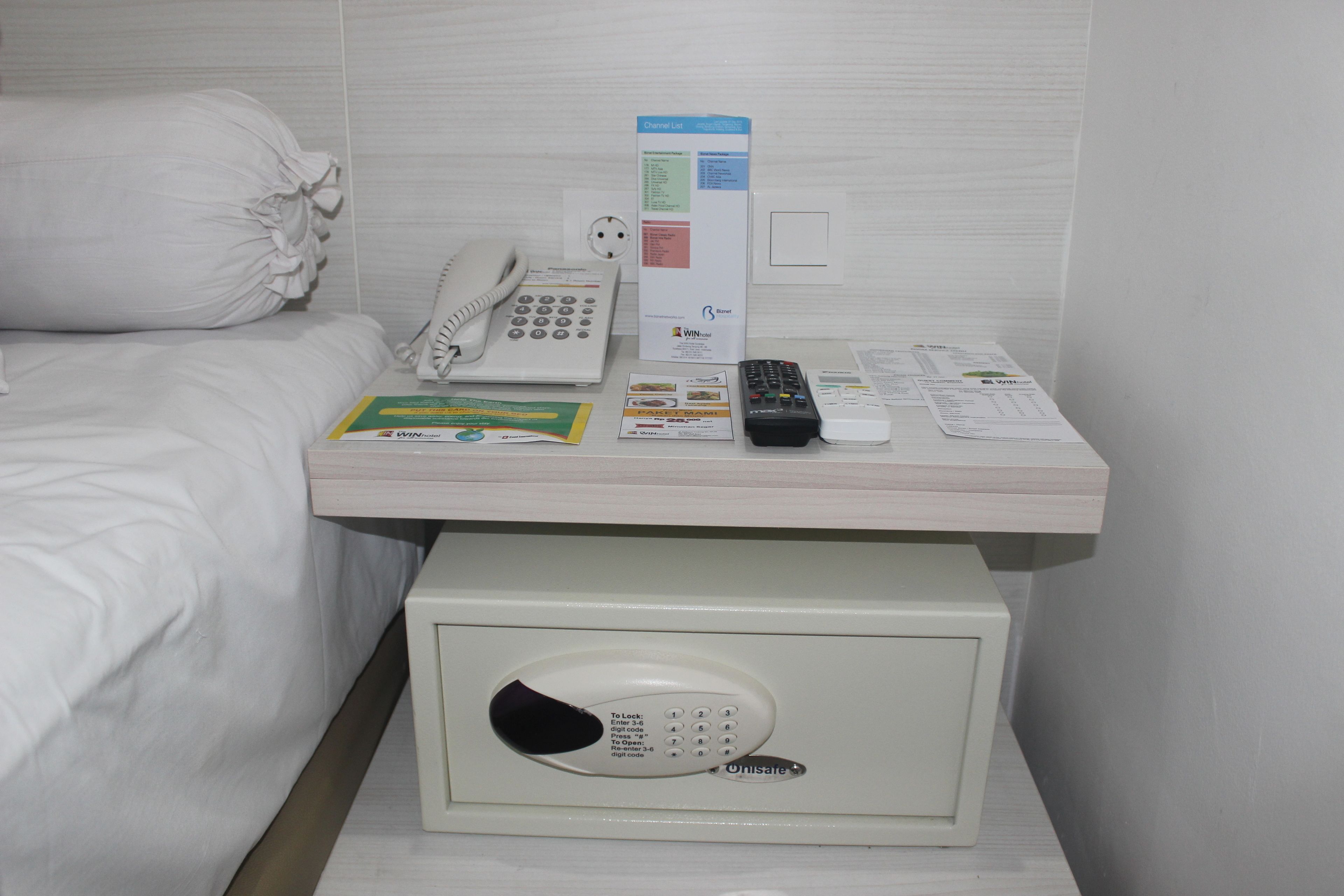 Habitación de hotel con mesita de noche que contiene un teléfono, un control remoto, un folleto y una caja de seguridad pequeña.